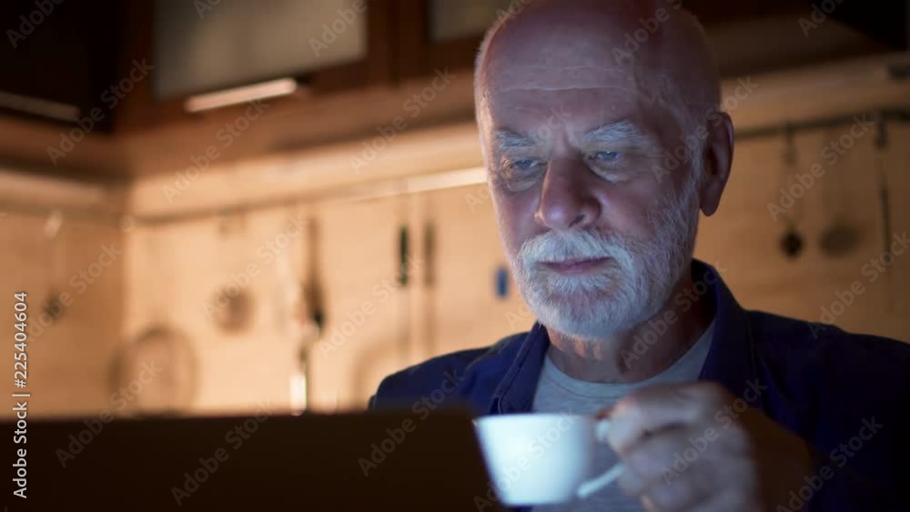 Senior man drink coffee from cup while read news on laptop. Male freelancer use computer at night work from home office. Overworked businessman work on project in kitchen in evening Senior man drink coffee from cup while read news on laptop. Male freelancer use computer at night work from home office. Overworked businessman work on project in kitchen in evening