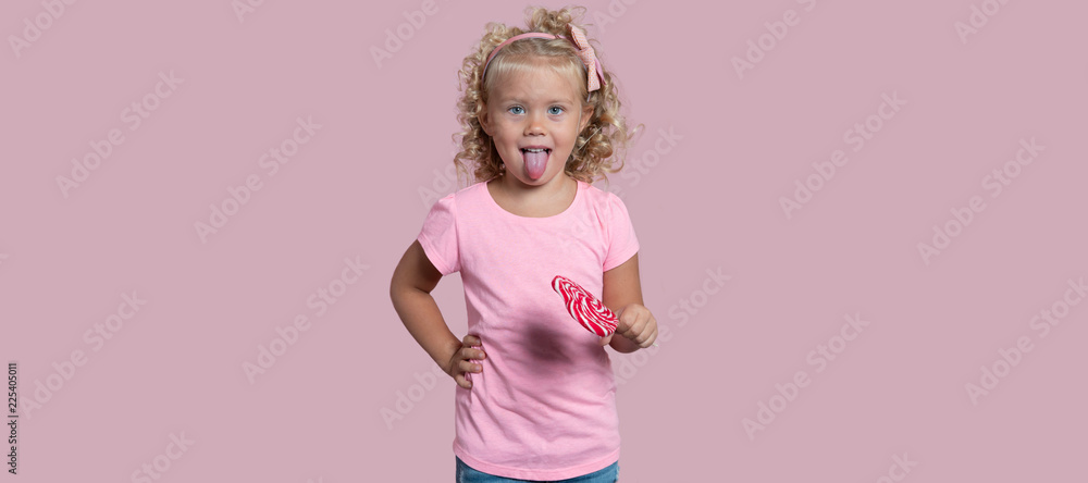 Fototapeta premium Small blond girl holds lollipop and shows a tong isolated over pink background