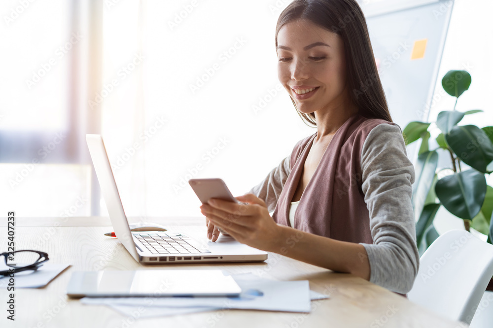 Message to business partner. Cheerful beautiful woman using her smartphone with smile at her working place. Message to business partner. Cheerful beautiful woman using her smartphone with smile at her working place.