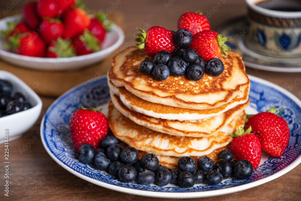Stack of pancakes with blueberries and strawberries on a blue and white plate with bowls of strawberries and blueberries and a cup of coffee in the background. 