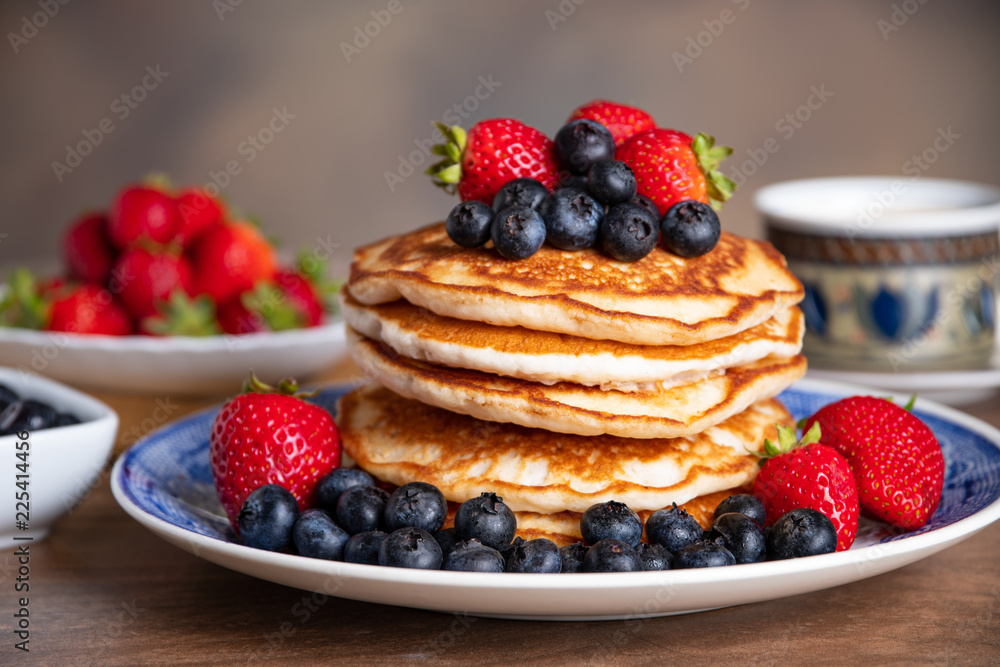 Stack of pancakes with blueberries and strawberries on a blue and white plate with bowls of strawberries and blueberries and a cup of coffee in the background. Stack of pancakes with blueberries and strawberries on a blue and white plate with bowls of strawberries and blueberries and a cup of coffee in the background.