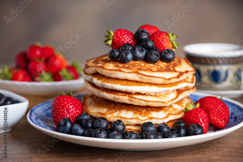 Stack of pancakes with blueberries and strawberries on a blue and white plate with bowls of strawberries and blueberries and a cup of coffee in the background.  photo