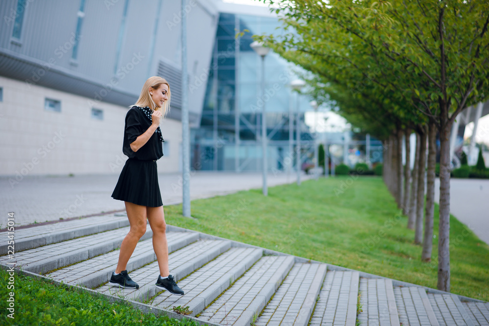 Handsome young blonde woman in black dress walking down the stairs near the office building outdoors on the street.