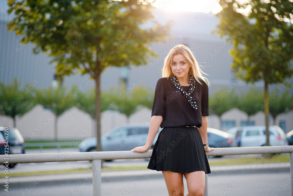 Attractive young blonde woman in stylish black blouse and skirt posing on the street near the road on sunset.