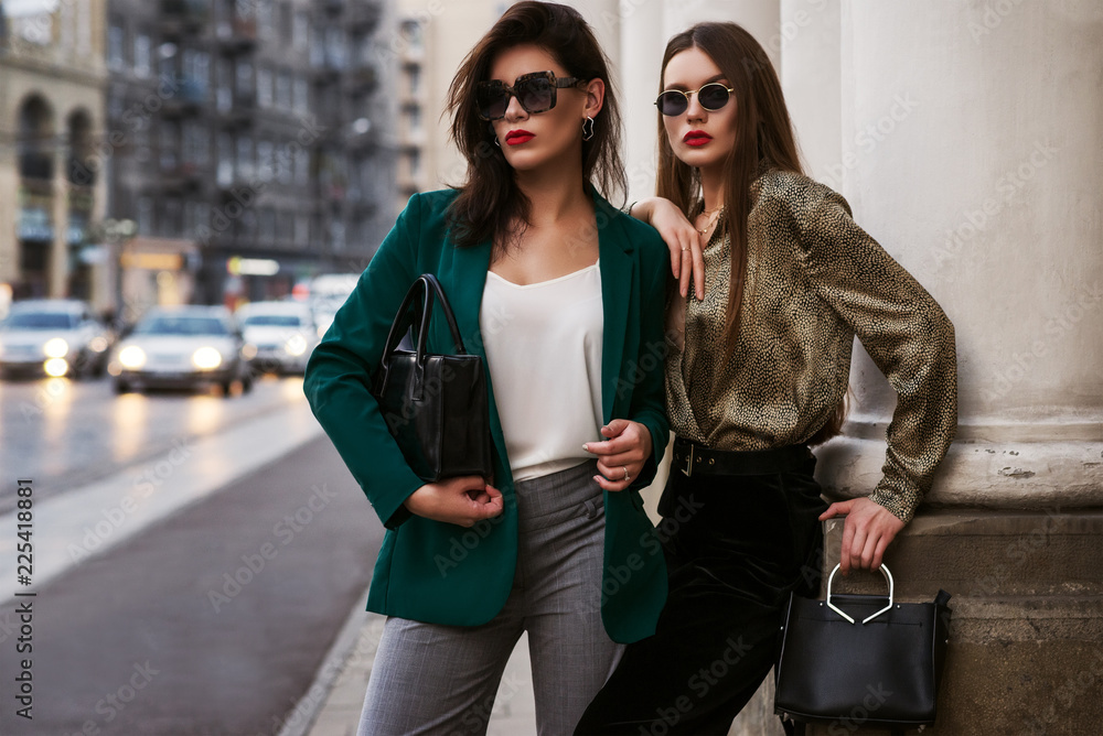 Outdoor fashion portrait of two young beautiful women wearing trendy sunglasses, stylish clothes, holding black leather bags, posing in street of european city. Copy, empty space for text - obrazy, fototapety, plakaty 