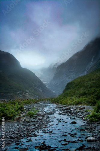 Glacial meltwater stream leading into the misty valley of Franz Josef Glacier on the South Island of New Zealand under a dramatic cloudy sky