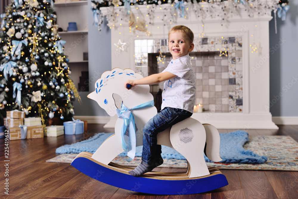 cute baby boy on a toy horse against the background of a Christmas tree - obrazy, fototapety, plakaty 