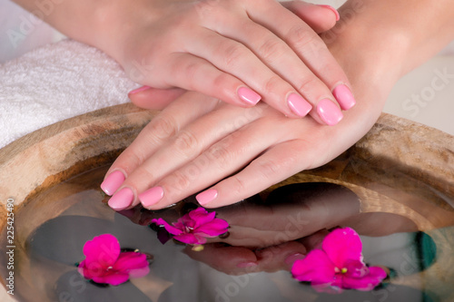 Gently woman hands with pink nails gel polish above water with purple flowers and black stones in wooden bowl. Manicure and beauty concept. Close up  selective focus