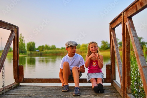Children sitting on pier. Two children of different age - elementary age boy and preschool girl sitting on a wooden pier. Girl making heart shape. Summer and childhood concept.  © romannerud