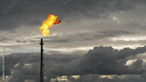 A tall GAS TORCH burns at an oil facility during dusk. The bright flame contrasts dramatically with the dark evening sky.