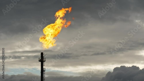 A tall GAS TORCH flares against the evening sky at an oil facility. Bright flame and dark clouds create a strong contrast.