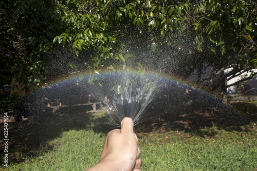 A hand is holding a garden rubber hose and spraying water on a green lawn and trees in bright sunlight, creating a rainbow in the process