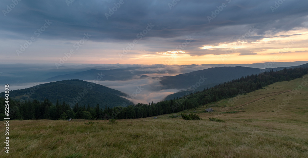 Landscape of mountain pasture, evening cloudy sky and green hillsides. Nature panoramic landscape. Carpathians mountains in august. Evening sun illuminating dense clouds. Blurred background
