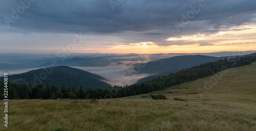 Landscape of mountain pasture, evening cloudy sky and green hillsides. Nature panoramic landscape. Carpathians mountains in august. Evening sun illuminating dense clouds. Blurred background