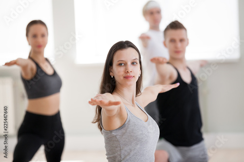 Group of young sporty people practicing yoga lesson, doing Warrior two exercise, Virabhadrasana II pose, working out, indoor, students training in club, studio close up. Well-being concept