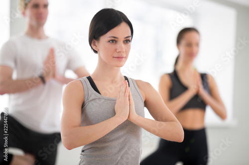 Portrait of young sporty attractive woman practicing yoga with group, doing meditation exercise vrikshasana pose with namaste, working out, indoor close up, yoga studio. Well-being concept