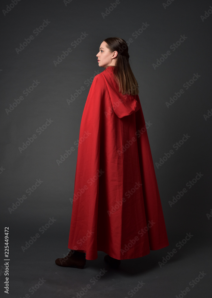 full length portrait of brunette girl wearing red medieval costume and cloak. standing pose  with back to the camera on grey studio background.