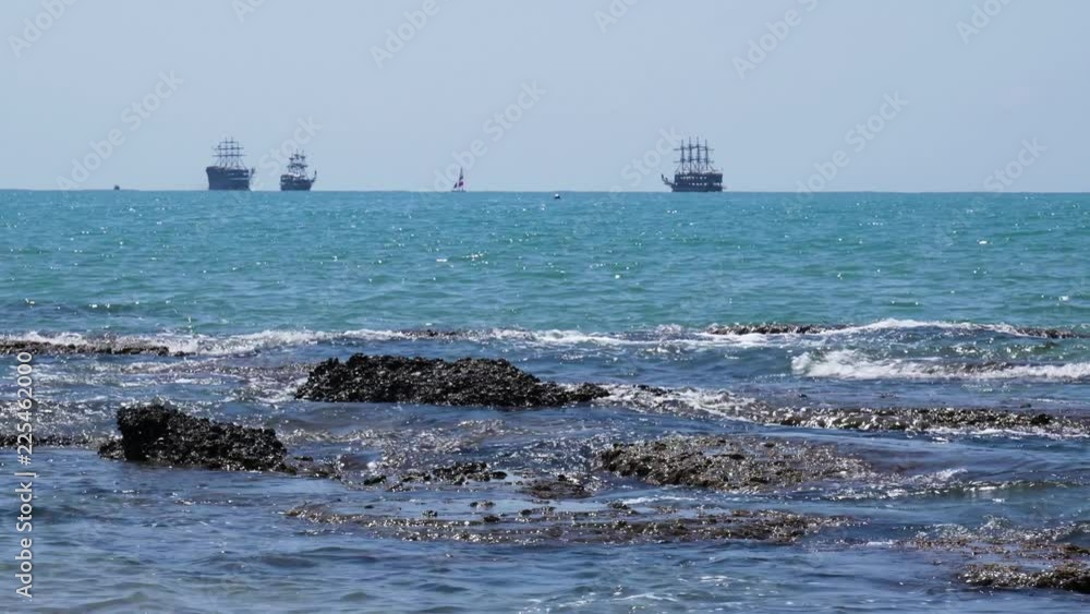 Three pirate ships sailing on horizon of blue ocean as waves break on rocky shore Three pirate ships sailing on horizon of blue ocean as waves break on rocky shore