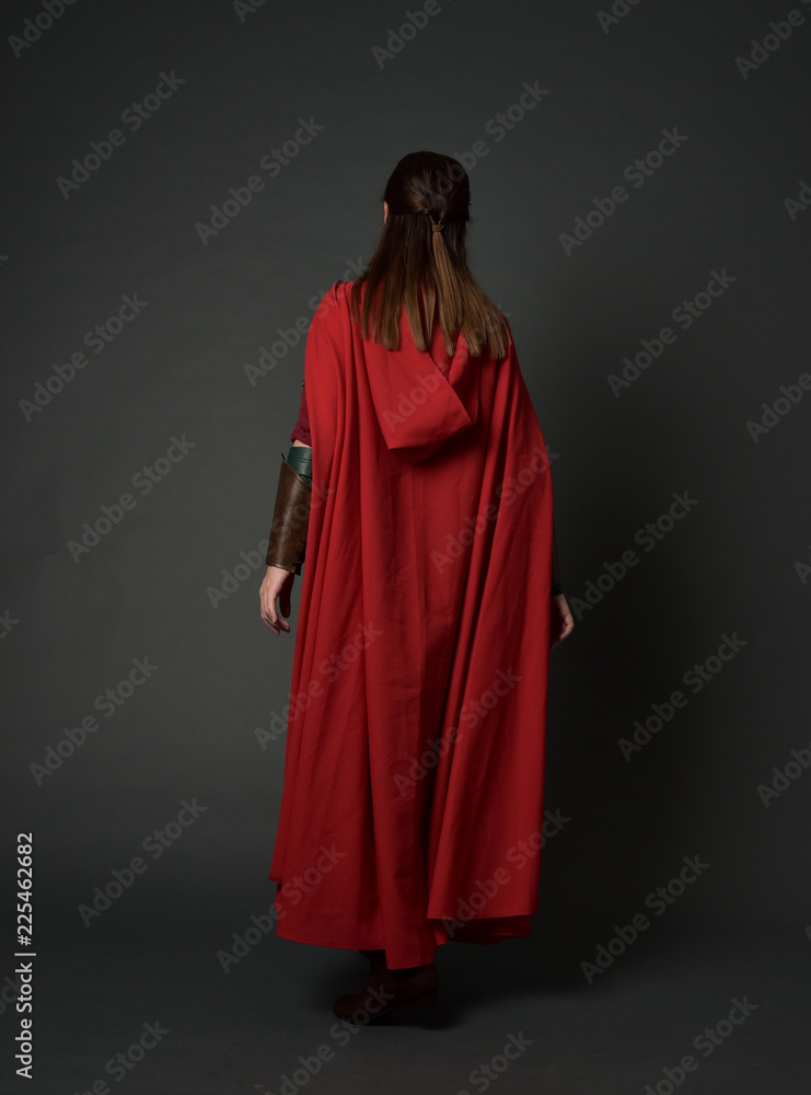 full length portrait of brunette girl wearing red medieval costume and cloak. standing pose  with back to the camera on grey studio background.