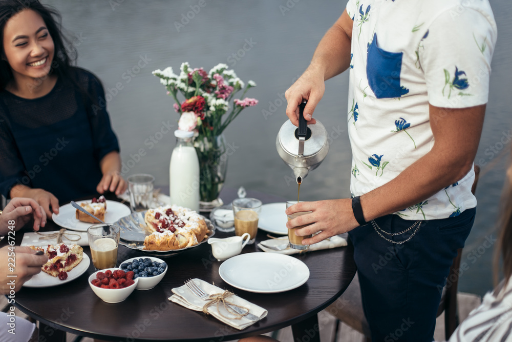 Young man with teapot pouring tea into cup during friendly breakfast Young man with teapot pouring tea into cup during friendly breakfast