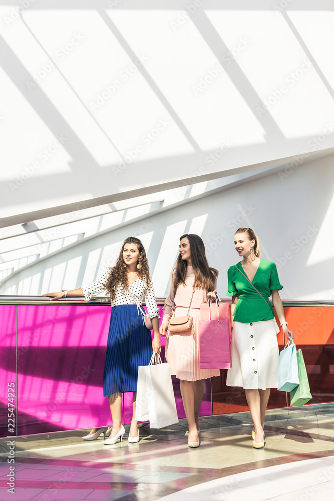 smiling fashionable girls holding paper bags and looking away at shopping mall smiling fashionable girls holding paper bags and looking away at shopping mall