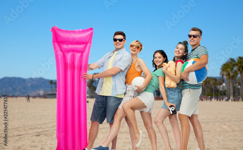 summer holidays, leisure and people concept - group of happy smiling friends in sunglasses with ball, volleyball, towel, camera and floating mattress over venice beach background in california