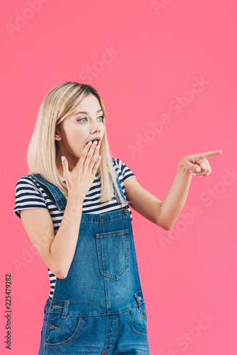 shocked girl in denim overall pointing on something and covering mouth isolated on pink