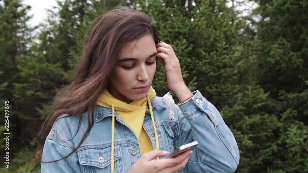 Young teen coed using touch screen smartphone while in nature. Hipster using social media and smiling while reading mobile. Beautiful woman in denim jacket in spruce forest Young teen coed using touch screen smartphone while in nature. Hipster using social media and smiling while reading mobile. Beautiful woman in denim jacket in spruce forest