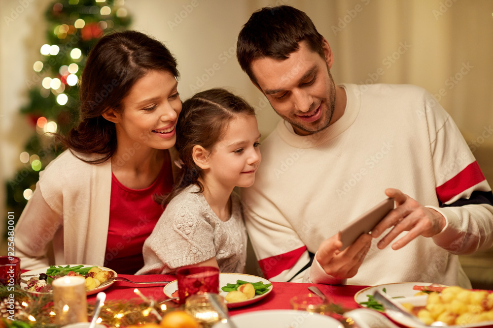 holidays, family and technology concept - happy mother, father and little daughter with smartphone having christmas dinner at home holidays, family and technology concept - happy mother, father and little daughter with smartphone having christmas dinner at home
