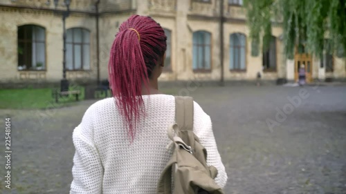 Moving shot of young african female student going to university, charming woman with backpack and pink dreadlocks