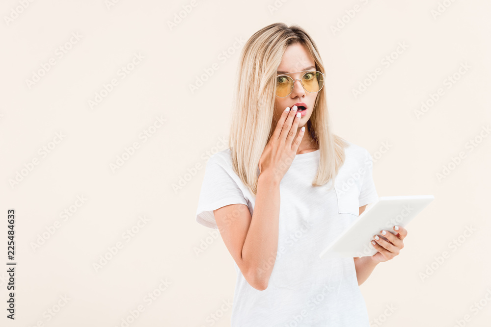 shocked stylish woman in eyeglasses covering mouth by hand and holding digital tablet isolated on beige shocked stylish woman in eyeglasses covering mouth by hand and holding digital tablet isolated on beige