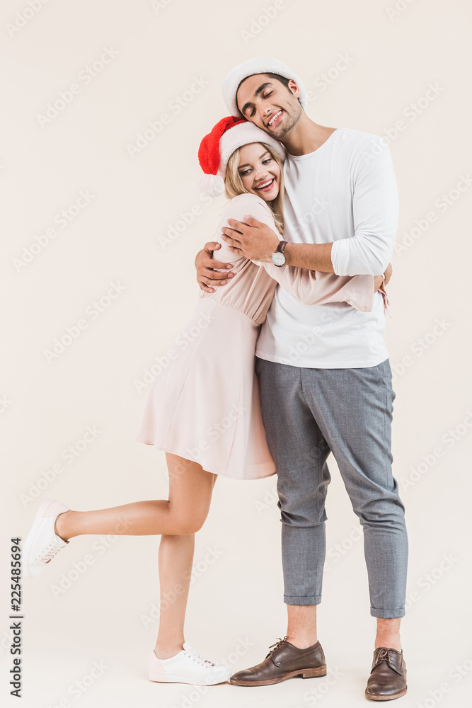 Obraz premium happy young couple in santa hats hugging isolated on beige