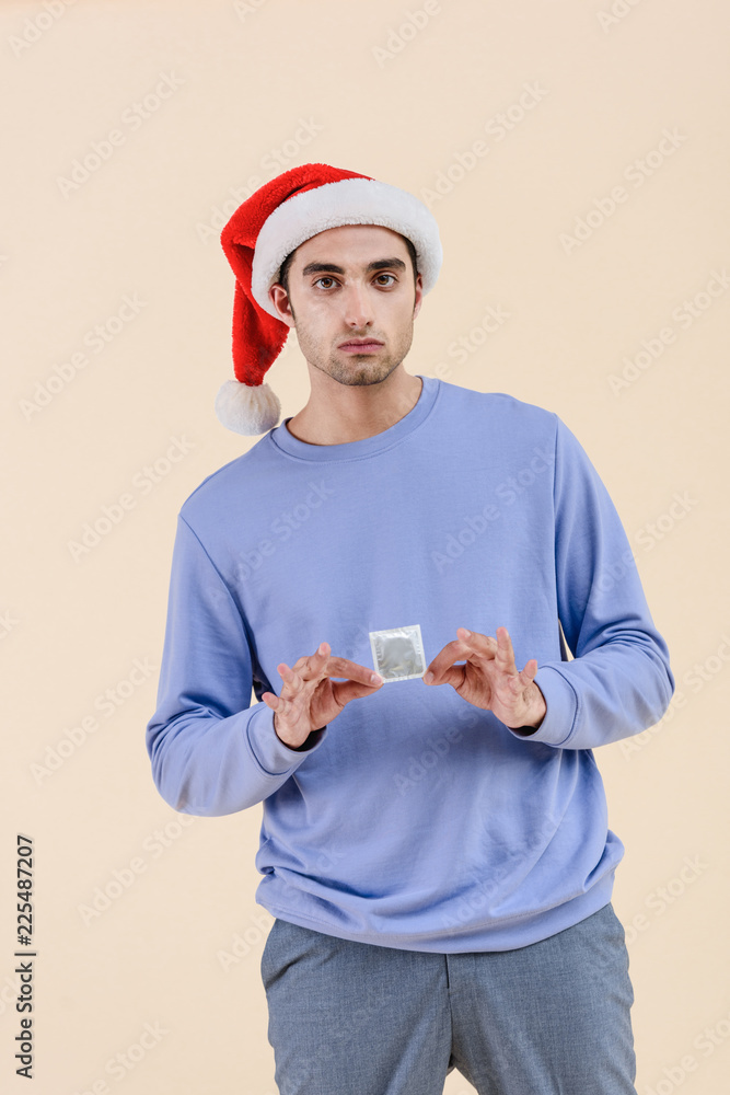 handsome young man in santa hat holding condom and looking at camera isolated on beige handsome young man in santa hat holding condom and looking at camera isolated on beige