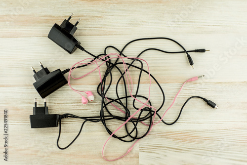the need for universal battery charger: black 3 different chargers with different connectors and earphones tangled cables together, on the white oak table