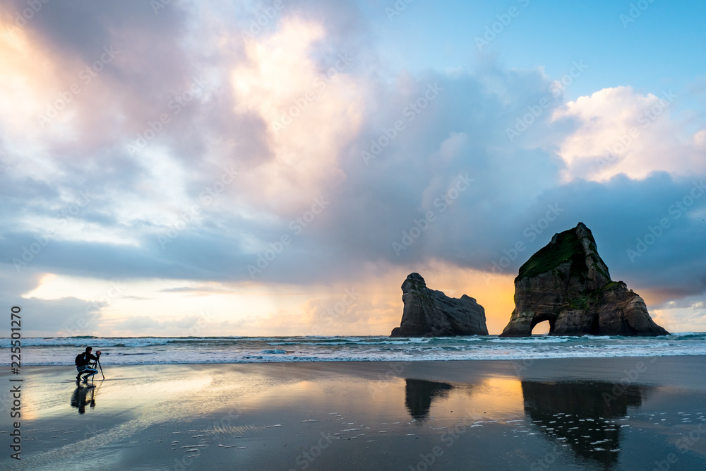 A photographer with the beautiful Wharariki Beach with famous rocks. Sunset scene golden light and silhouette. Nelson, South Island, New Zealand.
