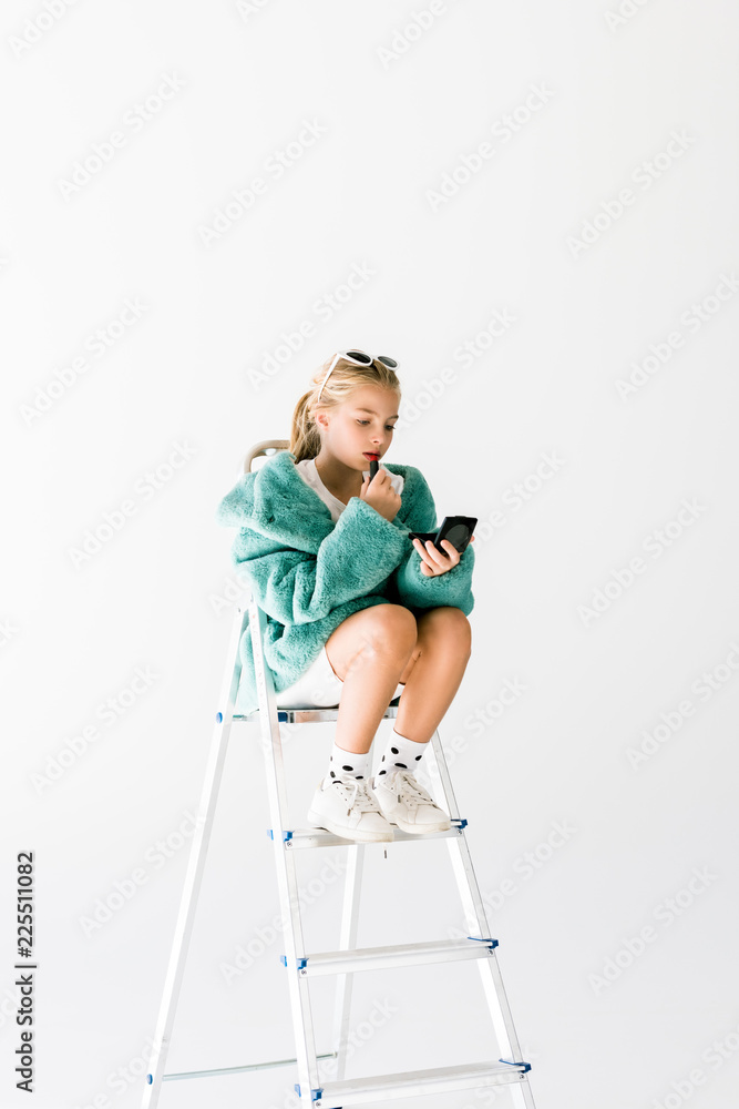 stylish kid in trendy fur coat applying lipstick and making makeup while sitting on ladder isolated on white