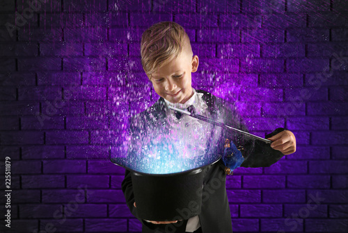 Cute little magician showing trick with hat against dark brick wall