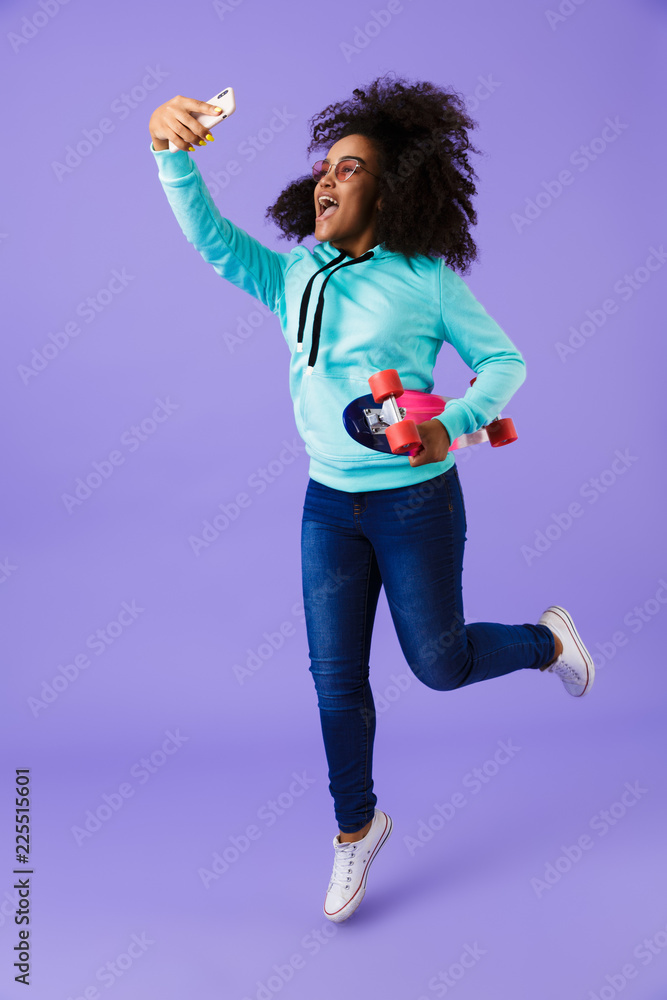 African girl posing isolated over violet background holding skateboard take a selfie by phone.