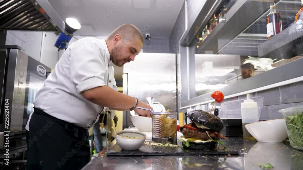 Cook in restaurant kitchen placing fried potatoes next to burger to serve clients Cook in restaurant kitchen placing fried potatoes next to burger to serve clients