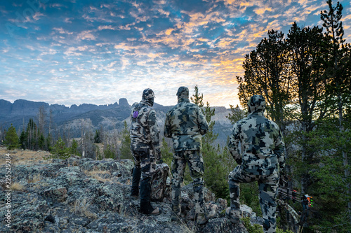 Tablou pe pânză Three adult male hunter friends, unrecognizable,  stand on a mountain ridge looking for elk to hunt during bow archery season