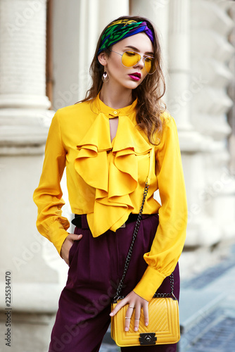 Outdoor fashion portrait of young fashionable woman wearing stylish headband, trendy colorful sunglasses, vintage yellow blouse with frills, violet trousers, holding small bag, posing in street