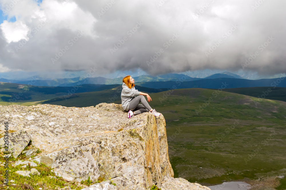 The beautiful red-haired girl sits and relaxing keep calm alone on the edge of a cliff in the rays of light on a rock Altai mountains on a background of blue sky with big gray clouds and landscape
