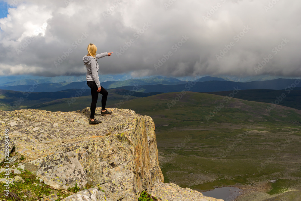 A young blond woman in a gray hoody, black leggins stands on a top of a cliff in the mountains of Altai and showing to the horizon by her arm with a lot of stones with picturesque landscape