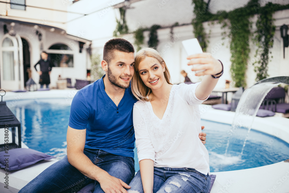 Young and attractive man and woman enjoying in private cocktail house party. They sitting, smiling and taking selfie photo. Swimming pool in background.
