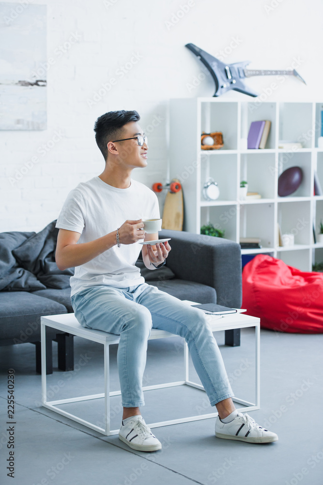 Φωτογραφία handsome smiling asian man sitting on table with cup of coffee in morning at hom