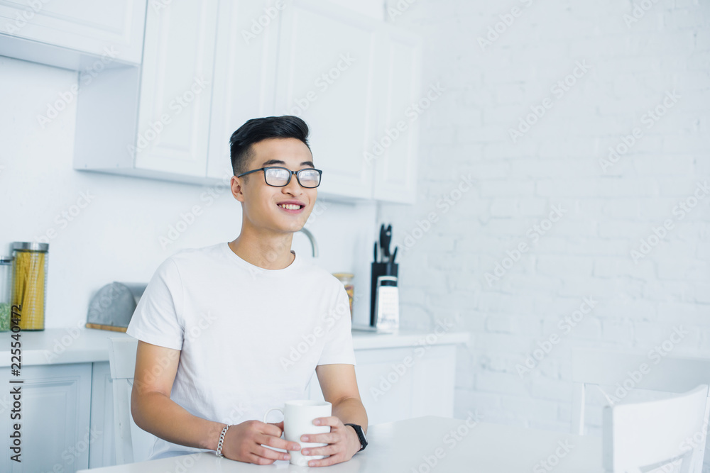 handsome smiling young asian man in eyeglasses holding cup of tea and looking away while sitting in kitchen handsome smiling young asian man in eyeglasses holding cup of tea and looking away while sitting in kitchen