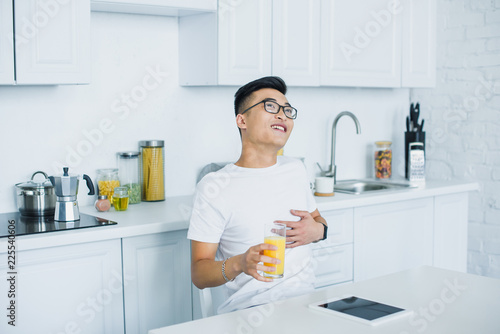 happy young asian man holding glass of juice and looking up while sitting in kitchen