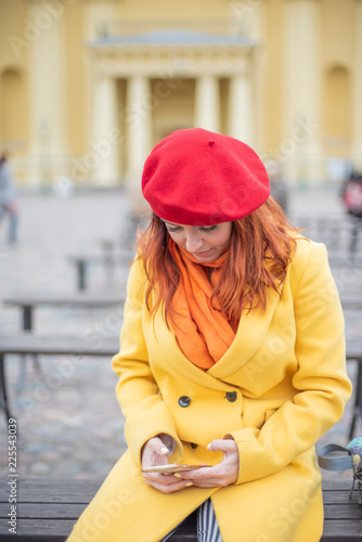 A red-haired young woman in a yellow coat and a red beret sits alone on a bench and looks into her smartphone. The girl sits alone on a bench and reads messages in the phone.