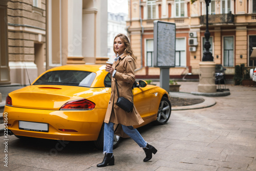 Pretty girl in trench coat with little black cross bag holding cup of coffee to go in hand dreamily looking in camera with yellow sport car on background on street © Anton