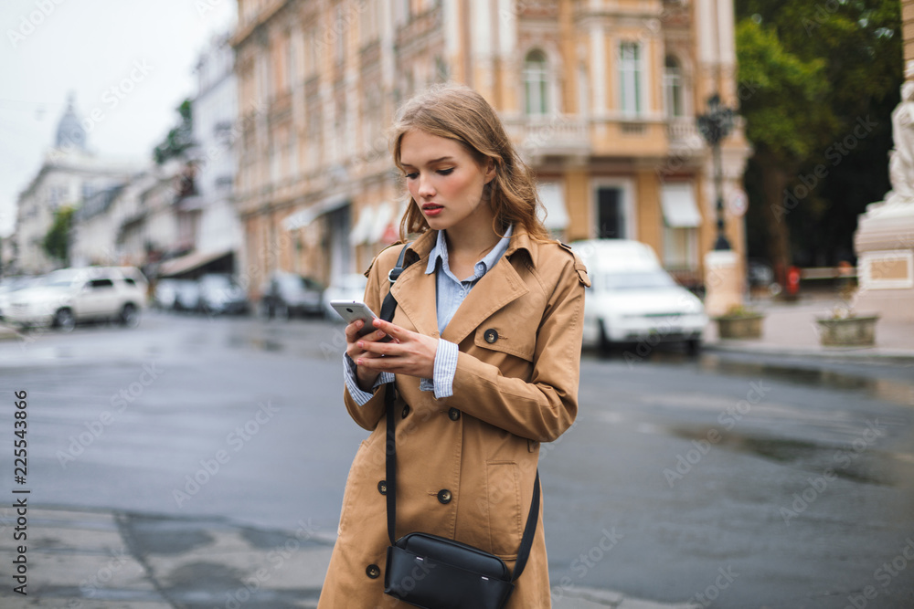 Beautiful girl in trench coat with little black cross bag thoughtfully using cellphone while spending time on cozy city street